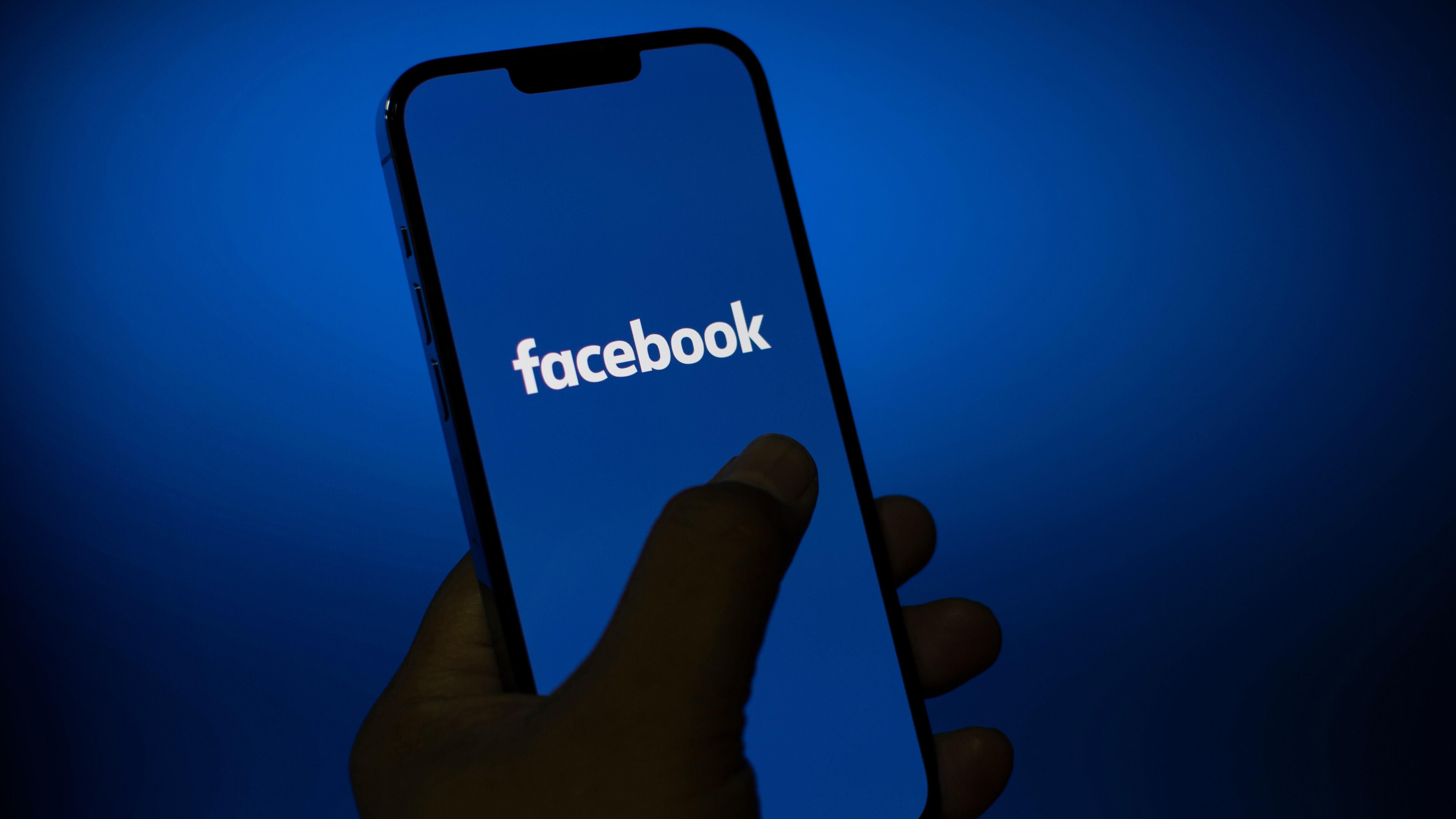 Facebook application on a mobile phone screen in hand, Social media platform on a smartphone on a blue background.