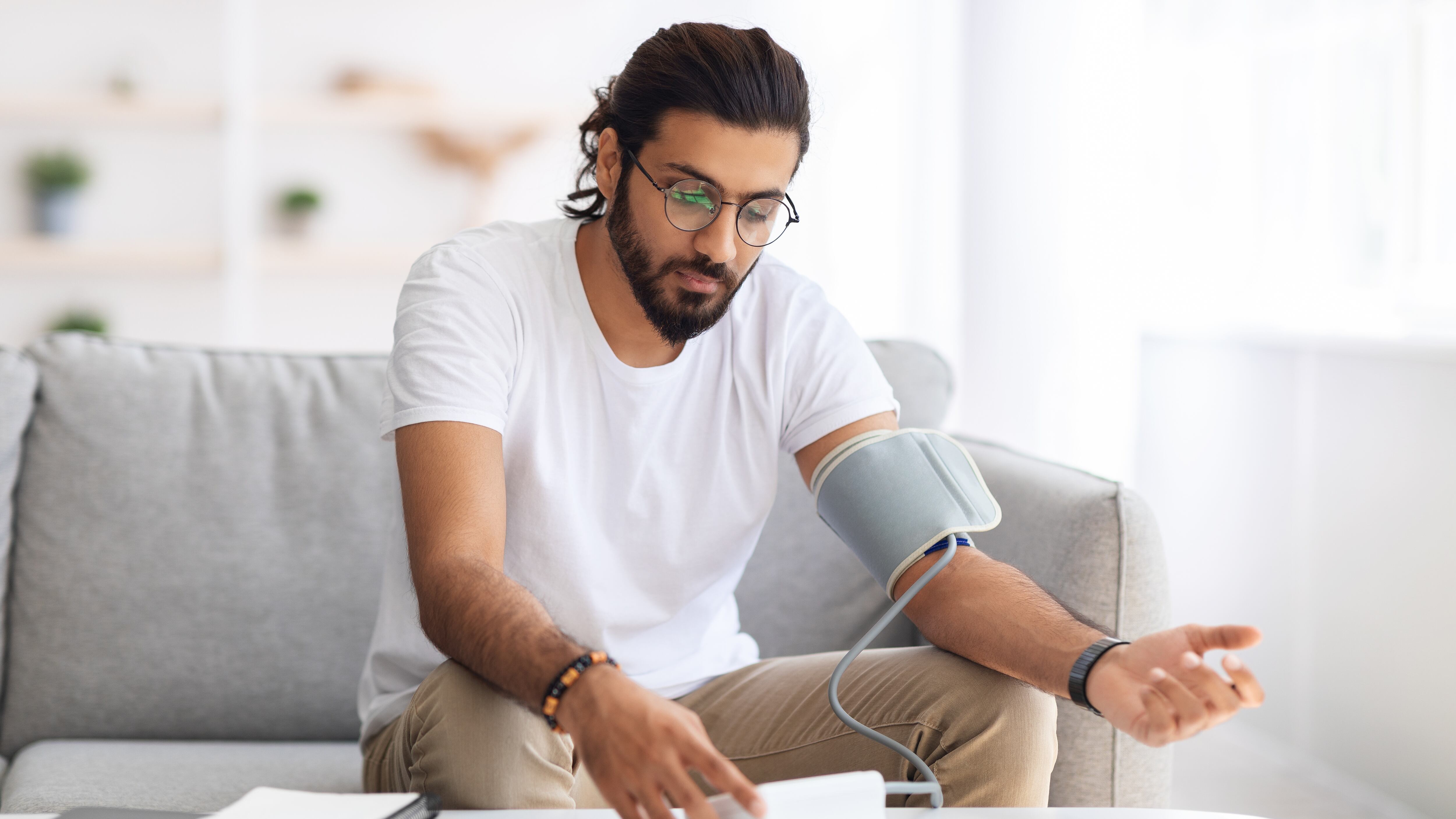 Young man sitting on couch, checking blood pressure and taking notes at home.